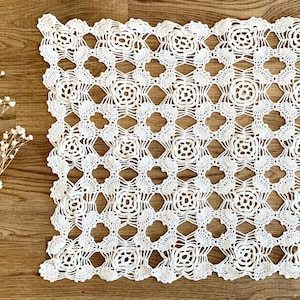 May include: White crocheted doily with a floral pattern. The doily is made of delicate, intricate stitches and has a square shape.