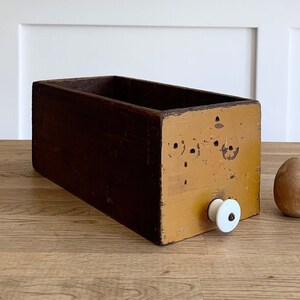 May include: A vintage wooden box with a yellow painted front and a white knob. The box has a worn, distressed look and is perfect for storage or display.