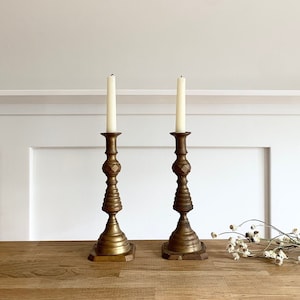 May include: A pair of gold metal candle holders with a geometric design. The candle holders are on a wooden surface with dried flowers.