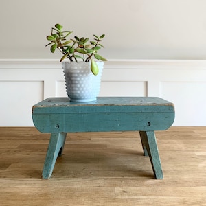 May include: A small, teal blue wooden stool with a distressed finish. The stool has a simple design with two legs on each side. A white milk glass planter with a succulent plant is sitting on top of the stool.