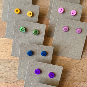 May include: Assortment of colourful button earrings displayed on small cards. The earrings are arranged in rows, with each row featuring a different colour. The colours include red, yellow, green, blue, purple, and black.