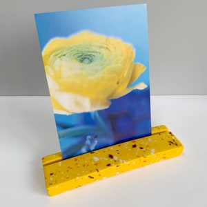 May include: A yellow wooden card holder with a speckled finish. A card with a yellow flower on a blue background is displayed in the holder.