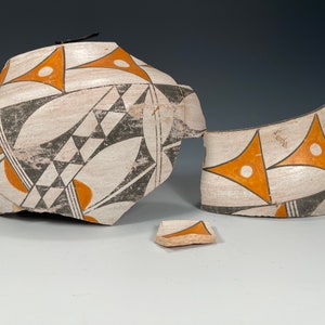 Puede incluir: Una olla de cerámica blanca rota con diseños geométricos negros y naranjas. La olla está en tres piezas.