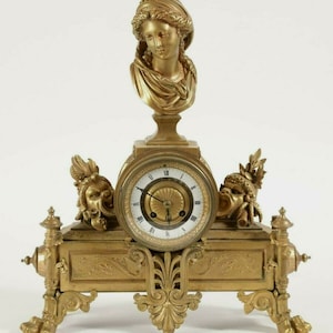 May include: A gold-toned mantel clock with a bust of a woman on top. The clock face has Roman numerals and a decorative centre. The clock is on a base with ornate details and two stylised faces on either side of the clock face.