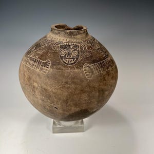 May include: A brown ceramic pot with a carved human face design. The pot has a rounded shape and a wide opening.