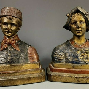 Puede incluir: Un par de sujetalibros vintage que representan a un niño y una niña con ropa tradicional. El niño lleva un sombrero rojo y una bufanda roja, mientras que la niña lleva un vestido azul y dorado y un sombrero blanco. Los sujetalibros están hechos de metal pintado y tienen un acabado dorado.