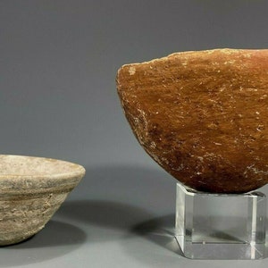 Può includere: Due ciotole antiche. La ciotola più piccola è grigio chiaro con striature orizzontali. La ciotola più grande è marrone con una consistenza ruvida e poggia su un supporto in acrilico trasparente. Le ciotole hanno una forma conica.