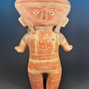 May include: A terracotta figurine of a person with a flat, wide head and a geometric design on the chest. The figurine is painted in shades of red and white.