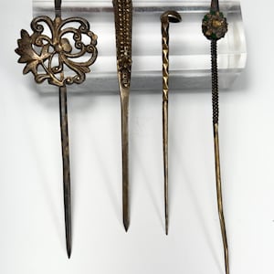 Op de afbeelding: Vier antieke gouden haarspelden met decoratieve details. De spelden zijn gemaakt van metaal en hebben verschillende ontwerpen, waaronder een bloemenmotief, een kralenontwerp, een geometrisch ontwerp en een bloemenontwerp met groen emaille.