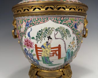 Könnte beinhalten: Eine chinesische Exportvase aus Porzellan und vergoldetem Bronze mit einem rosa-weißen Blumendesign und einer Figur in einer Gartenszene. Die Vase hat einen Deckel und steht auf einem dreifüßigen Sockel.