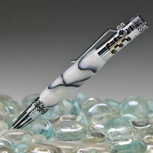 Może przedstawiać: Srebrny i biały długopis z czarno-białym wzorem marmurowym. Długopis ma srebrny klips i srebrny mechanizm skręcany. Długopis leży na łóżku z przezroczystych szklanych kamieni.