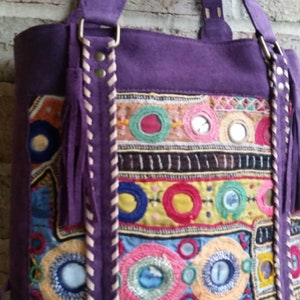 May include: Purple suede tote bag with colorful embroidered details and round mirrors. The bag has a long strap and a tassel.