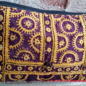 May include: A purple and gold embroidered clutch bag with a zipper closure and a chain strap. The bag is decorated with small round mirrors.