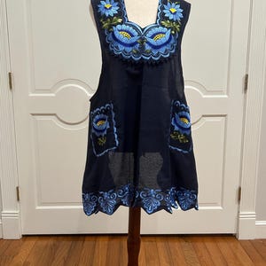 May include: A navy blue embroidered dress with blue floral designs. The dress features a square neckline and two pockets, with a scalloped hemline. The dress is displayed on a wooden dress form.