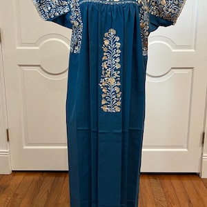 May include: A teal-colored, floor-length dress with short sleeves. The dress features intricate gold floral embroidery around the neckline and sleeves, and down the center. The dress is displayed on a mannequin.