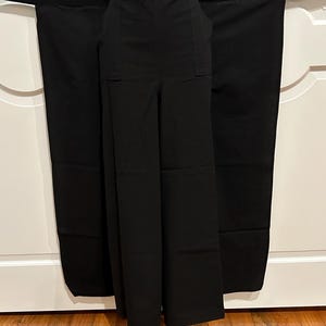 May include: Black wide-leg pants with two front pockets and a high waist. The pants are displayed on a mannequin with a matching black sash tied around the waist. The fabric appears to be a smooth, solid material.