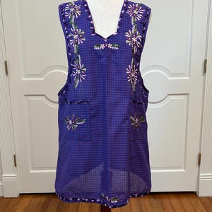 May include: A sleeveless, purple apron-style garment with a checkered pattern. The apron features embroidered floral designs in white, purple, and green. It has two pockets and a scalloped edge. The garment is displayed on a wooden mannequin.