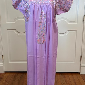 May include: A long, lavender dress with short sleeves and intricate floral embroidery on the bodice and sleeves. The dress is displayed on a mannequin, showcasing its flowing design and detailed craftsmanship. The dress is made of a soft, lightweight fabric.