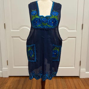 May include: A navy blue embroidered dress with blue and green floral designs. The sleeveless dress features a square neckline, two front pockets, and a scalloped hem. The dress is displayed on a wooden dress form.