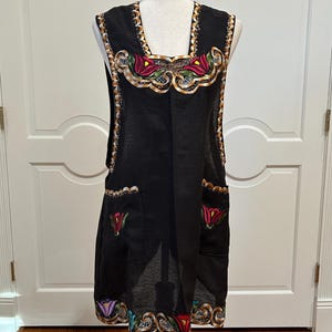 May include: Black apron with colorful floral embroidery. The apron features a decorative gold trim along the edges and around the embroidered details. Two pockets are visible on the front, and the apron is displayed on a mannequin.