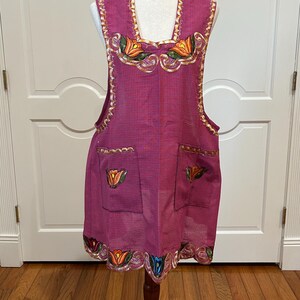 May include: A magenta apron with embroidered floral designs in orange, yellow, and green. The apron has two pockets and gold trim along the edges. It is displayed on a mannequin.