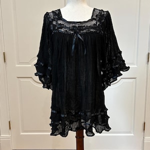 May include: A black tunic featuring a square neckline with a grid-like crochet pattern, a small central ribbon bow, and three-quarter length ruffled sleeves. The garment is made of a sheer, textured fabric with ribbon trim along the hem and sleeves.