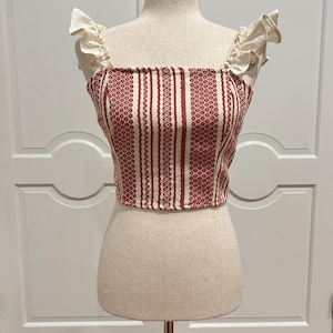 May include: A red and white striped crop top with ruffled off-white straps. The top is displayed on a mannequin. The fabric has a textured pattern. The top is a square neck style.