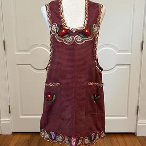 May include: A burgundy apron with a checkered pattern, adorned with floral embroidery in red, yellow, and green. The apron features scalloped edges and two pockets, displayed on a mannequin.