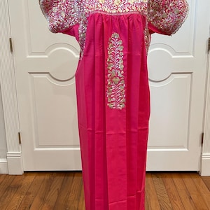 Puede incluir: Un vestido rosa brillante con mangas cortas y cuello redondo. Las mangas y el cuello están adornados con un estampado floral en dorado, rosa y blanco. Un bordado floral vertical en dorado y blanco adorna la parte delantera del vestido.
