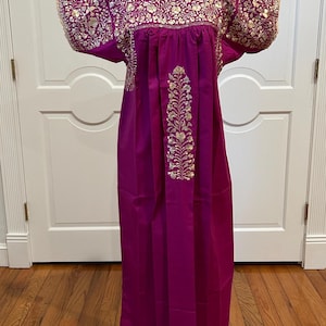May include: A vibrant magenta dress with intricate gold floral embroidery on the neckline and sleeves. The dress features a long, flowing silhouette with a central embroidered design. The dress is displayed on a mannequin.