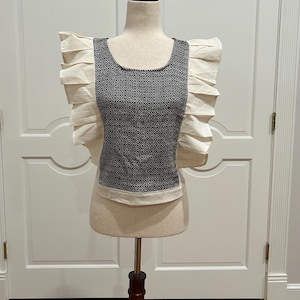 May include: A sleeveless top with a black and white geometric pattern on the front and cream-colored ruffled sleeves. The top is displayed on a mannequin with a wooden stand, set against a white door background.