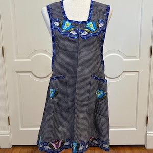May include: A sleeveless apron in a gray and white checkered pattern, adorned with blue floral embroidery. The apron features two pockets and decorative trim along the edges. The apron is displayed on a mannequin.