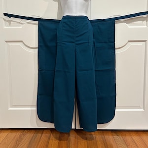 Puede incluir: Pantalones anchos de color verde azulado con cintura alta y dos bolsillos delanteros. Los pantalones tienen largos paneles de tela adjuntos que se envuelven alrededor de la cintura. La prenda se exhibe en un maniquí blanco.