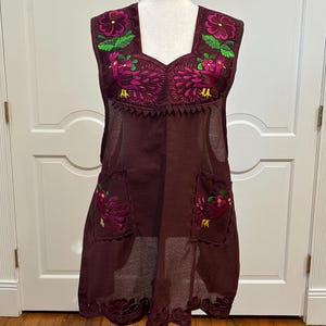 May include: A sleeveless burgundy apron-style garment with intricate floral embroidery in pink, green, and purple. The embroidery features bird and flower designs. The garment has two pockets and a scalloped hemline. The fabric appears to be semi-sheer.