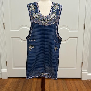 May include: A sleeveless blue tunic with a square neckline and embroidered floral details. The tunic has a vertical striped pattern and is displayed on a mannequin. The embroidery features white, green, and pink accents.