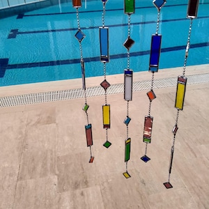 Peut inclure: Un ensemble de capteurs solaires en vitrail colorés suspendus devant une piscine. Les capteurs solaires présentent des pièces de verre rectangulaires et en forme de losange de différentes couleurs, notamment le bleu, le vert, l'orange et le jaune, reliées par des chaînes argentées.