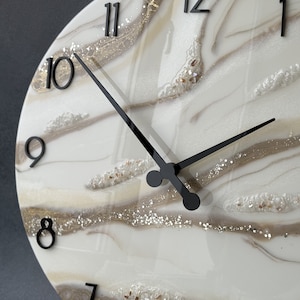 Puede incluir: Un reloj de pared redondo con un diseño veteado blanco y beige. La esfera del reloj presenta números y manecillas negras. El diseño incluye detalles de purpurina dorada y plateada, creando un aspecto decorativo y elegante.