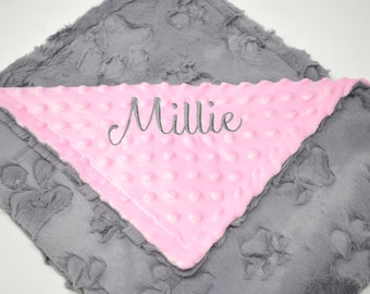 May include: A gray and pink baby blanket with the name "Millie" embroidered in gray script. The blanket has a textured, plush gray exterior and a smooth, pink interior with raised dots.