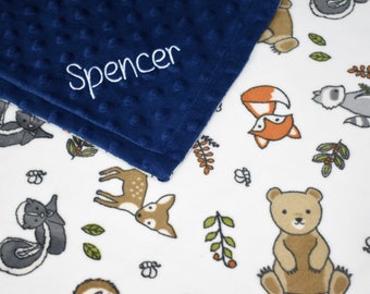 May include: A white baby blanket with a navy blue minky side. The blanket features a woodland animal print with deer, foxes, bears, and raccoons. The navy blue side has the name "Spencer" embroidered in white.