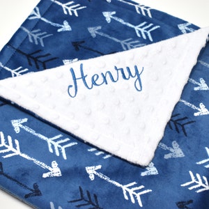 May include: A blue and white baby blanket with a white minky dot fabric on one side and a blue side with a white arrow pattern. The blanket is personalized with the name "Henry" embroidered on the white side.