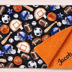May include: A black sports-themed blanket featuring basketballs, footballs, baseballs, baseball bats, basketball hoops, and blue football helmets. The blanket has an orange minky backing and the name "Jacob" in black script.