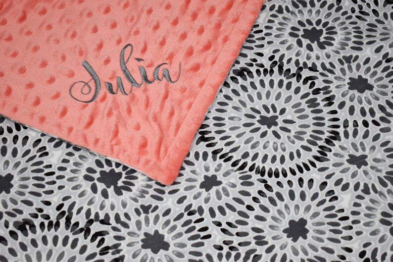 K&ouml;nnte beinhalten: Eine korallenrosa Minky-Babydecke mit einem schwarzen und wei&szlig;en Wirbelmuster auf der R&uuml;ckseite. Die Decke ist mit dem Namen "Julia" bestickt, der in grauer Farbe gestickt ist.