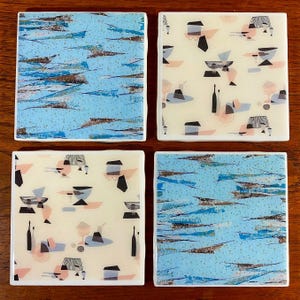 May include: Set of four square ceramic coasters with a blue and white abstract design. Two coasters feature a blue and white abstract design, and two coasters feature a white background with a repeating pattern of black and pink abstract shapes.