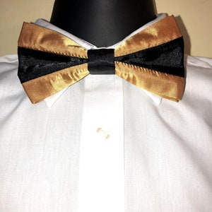 Puede incluir: Una pajarita dorada y negra. La pajarita está hecha de satén y tiene una franja negra en el medio.