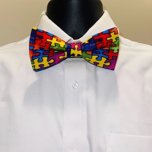 May include: A colorful bow tie featuring a jigsaw puzzle pattern in shades of red, yellow, green, blue, and purple. The bow tie is set against a white dress shirt, showcasing its vibrant design. This accessory adds a pop of color to any outfit.