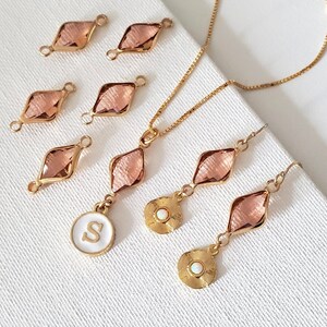 May include: A set of gold jewellery featuring a necklace, earrings, and charms. The necklace has a delicate chain with a pendant featuring a pink crystal and a gold disc with a white opal. The earrings are similar, with pink crystal drops and gold discs with white opals. The charms are small, with pink crystals and gold loops. The jewellery is on a white background.