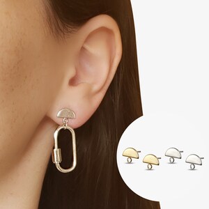 May include: Silver earrings with a half-circle top and a dangling oval link. The earrings are shown on a model's ear. The image also shows three pairs of earrings in gold, silver, and black.