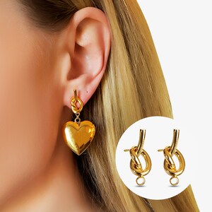 May include: A pair of gold-toned earrings featuring a polished heart-shaped locket pendant suspended from a knotted post. The design includes a smooth, reflective metallic finish on the heart charm and the knot detail, creating a cohesive look.