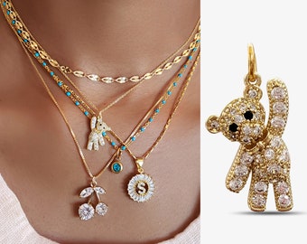 May include: A gold chain necklace with multiple pendants, including a cherry, a turquoise eye, a bunny, and a letter 'S' charm. The necklace is layered on a model's neck. The image also shows a separate gold pendant in the shape of a teddy bear with white cubic zirconia stones.