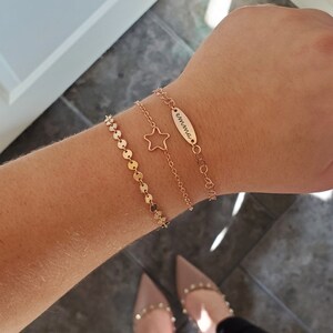 Puede incluir: Tres pulseras de oro apiladas en una mu&ntilde;eca. La pulsera superior es una cadena fina con una peque&ntilde;a etiqueta ovalada que dice "emma". La pulsera del medio es una cadena delicada con un dije de estrella. La pulsera inferior es una cadena de peque&ntilde;os c&iacute;rculos dorados.